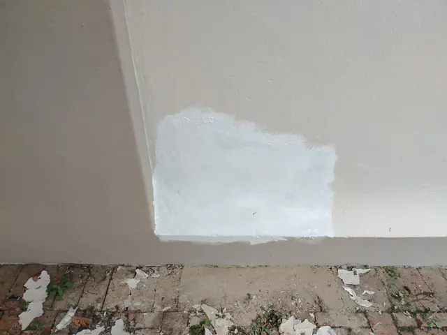 Beige exterior wall with a white primed patch and paint flakes on the paving below