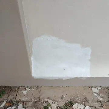Beige exterior wall with a white primed patch and paint flakes on the paving below